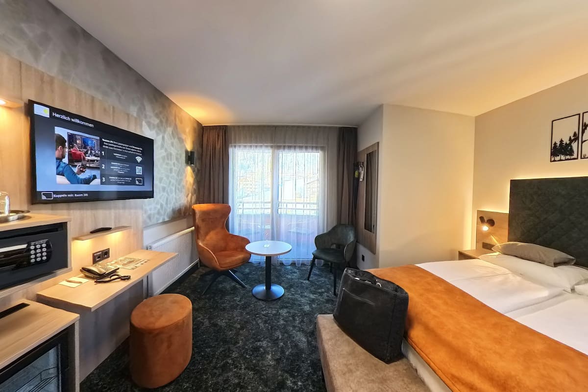 A hotel room with a bed, a desk with a TV, a chair, a small table, and a window with curtains.
