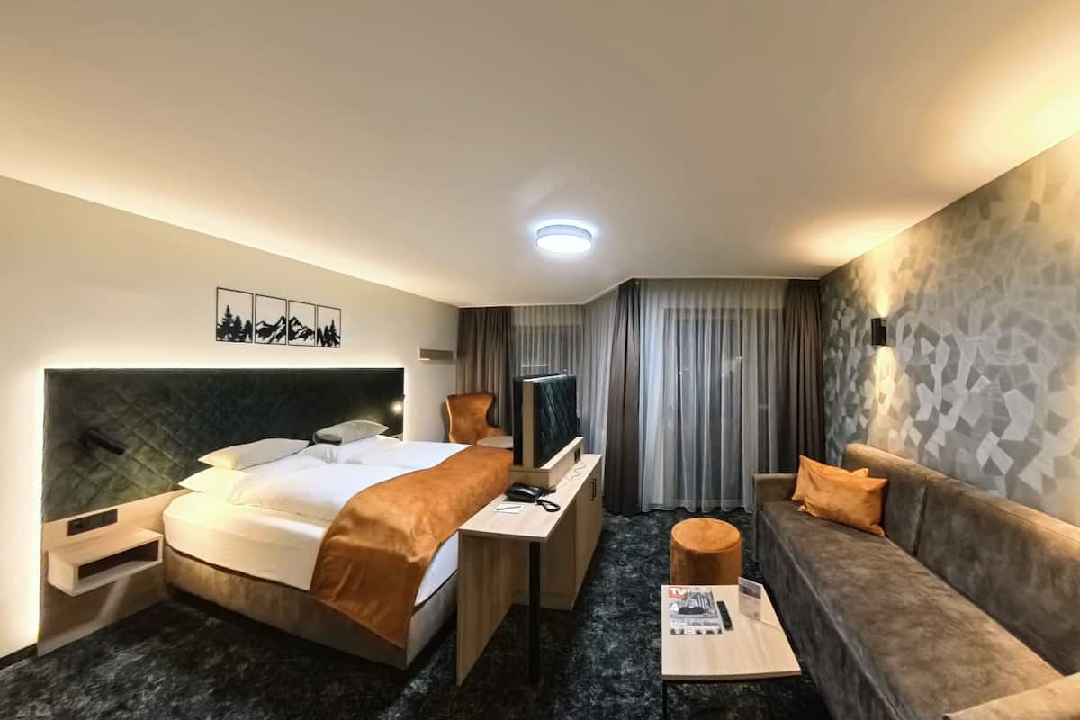 A hotel room with a bed, a sofa, a desk with a chair, a TV, and a wall-mounted light.