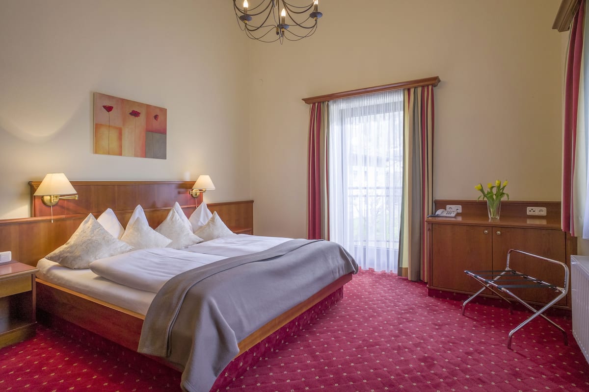 A hotel room with a large bed, two bedside lamps, a painting on the wall, a window with curtains, a radiator, and a small table with a vase of flowers.