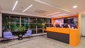 A reception area with a modern design, featuring a reception desk, two blue chairs, and a large window with plants. - Transamerica Fit Villa Lobos (São Paulo)