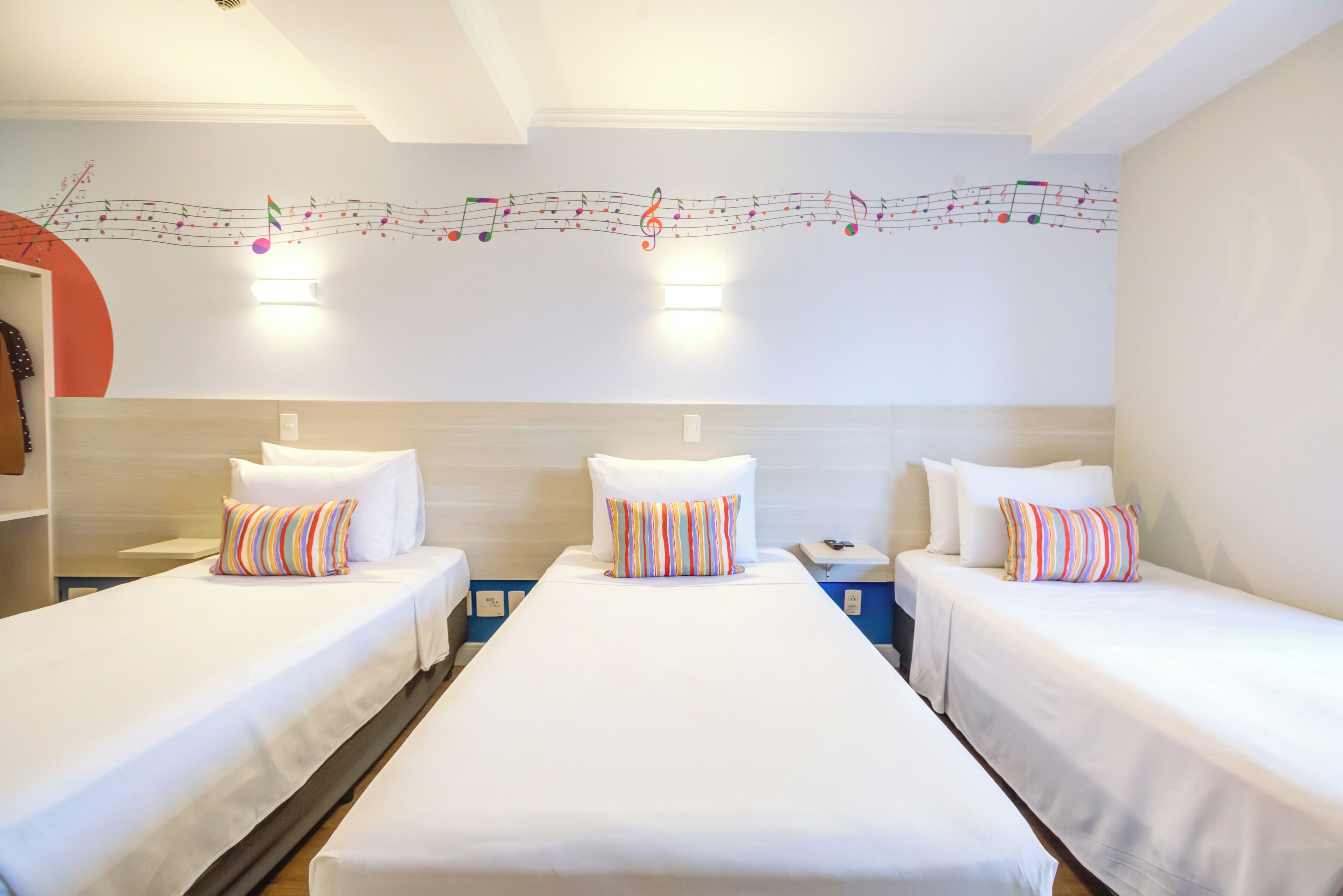 Quarto de hotel com três camas de solteiro, cada uma com roupa de cama branca e almofadas listradas. Há um candeeiro de parede sobre as camas e uma obra de arte decorativa com notas musicais.