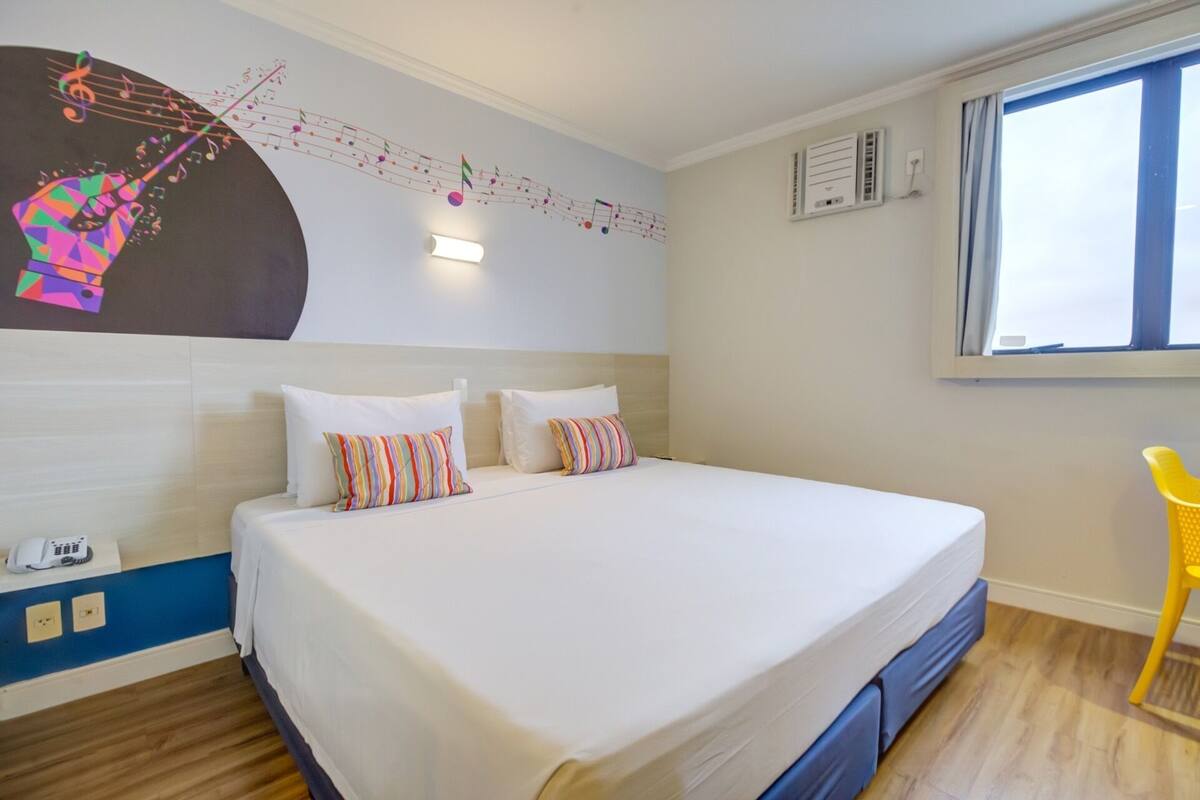 Ein Hotelzimmer mit einem Bett, zwei Kissen, einer Wanddekoration, einem Fenster und einem Sessel.