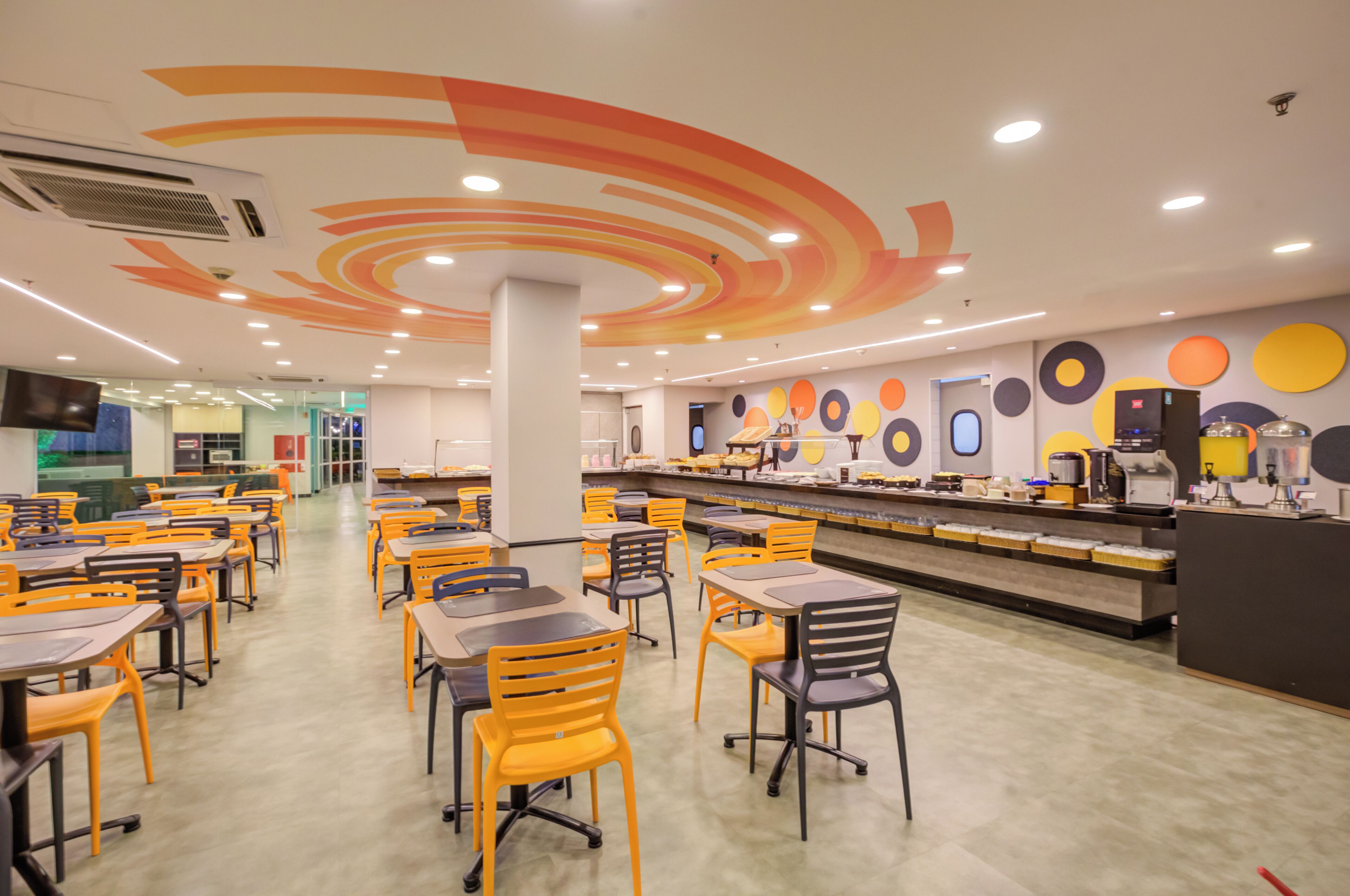 Eine moderne Cafeteria mit mehreren Tischen und Stühlen, einer langen Theke mit Speisenausgabe und einem farbenfrohen Deckenmuster.