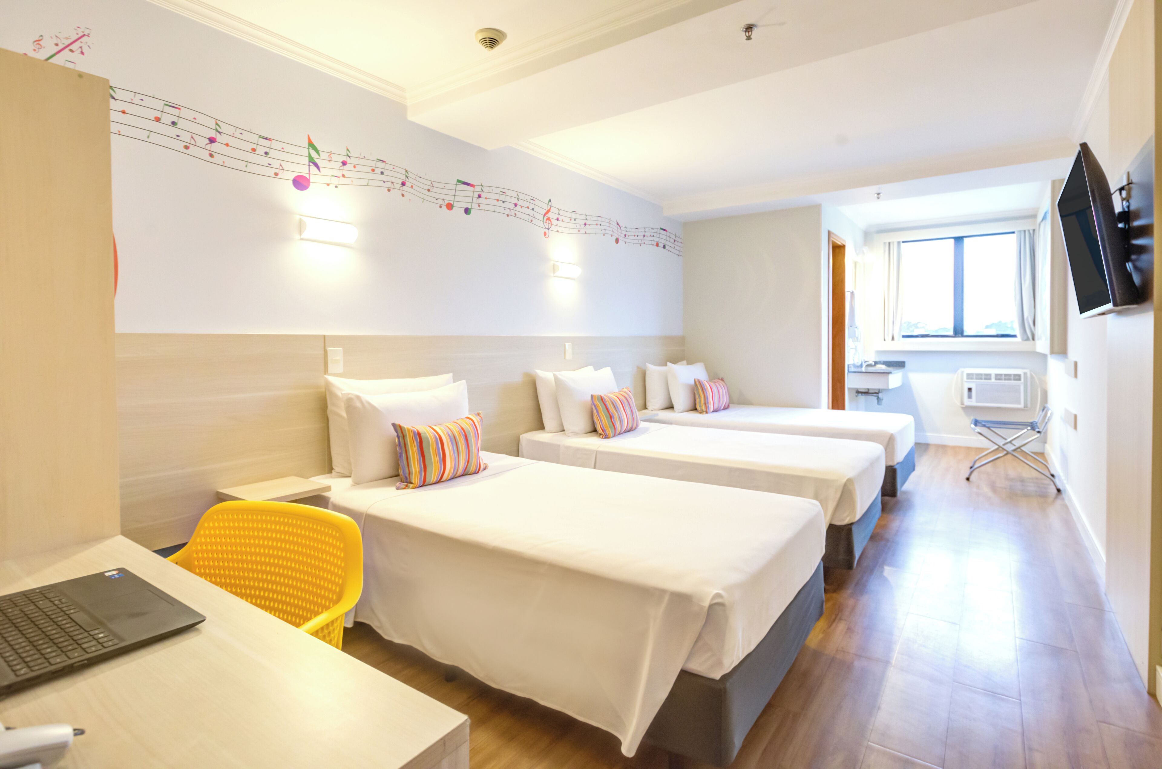 Une chambre d’hôtel avec trois lits, un bureau avec un ordinateur portable, une chaise et un téléviseur fixé au mur.