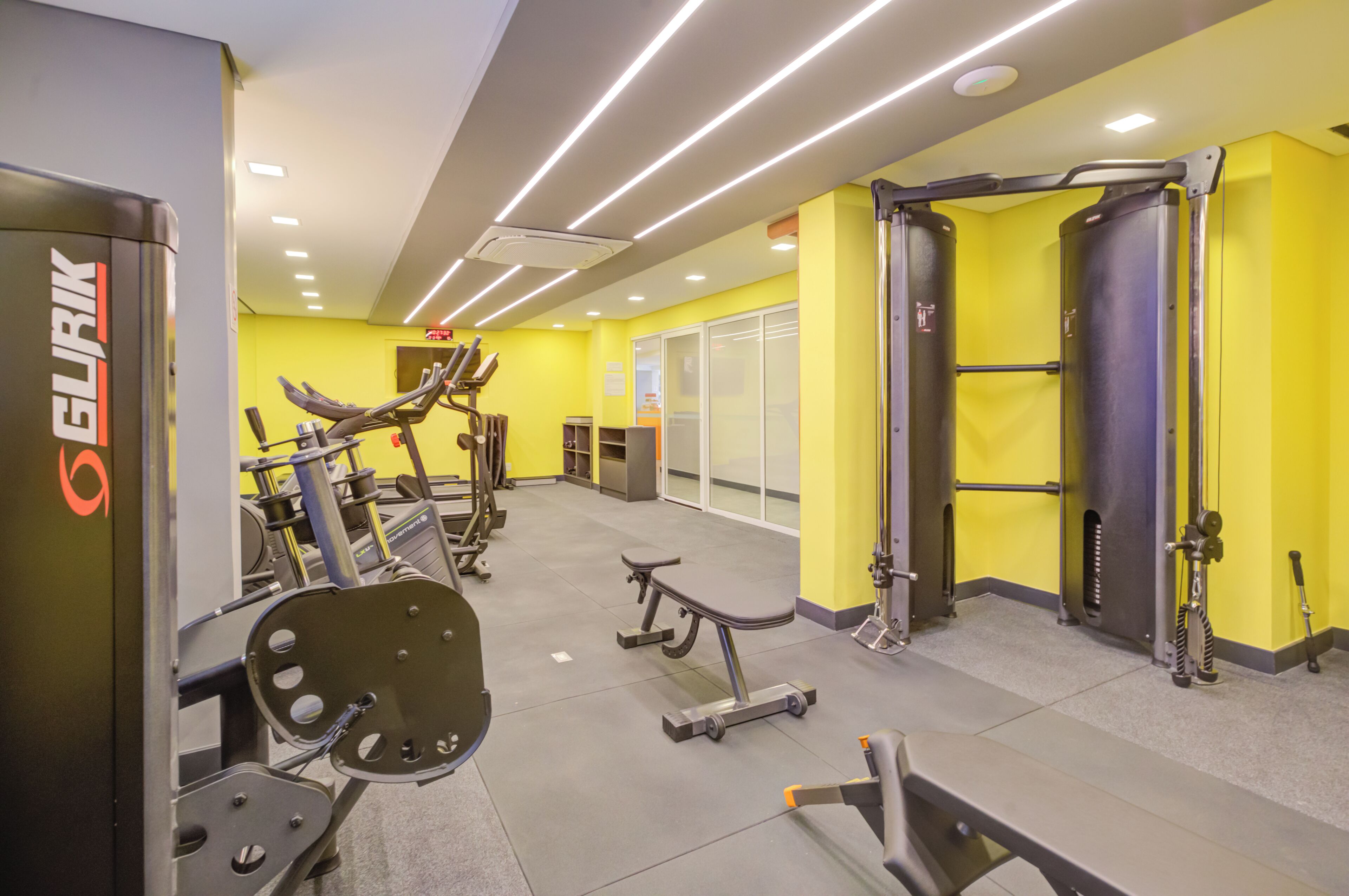 A gym with various exercise equipment, bright yellow walls, and a ceiling with linear lighting.