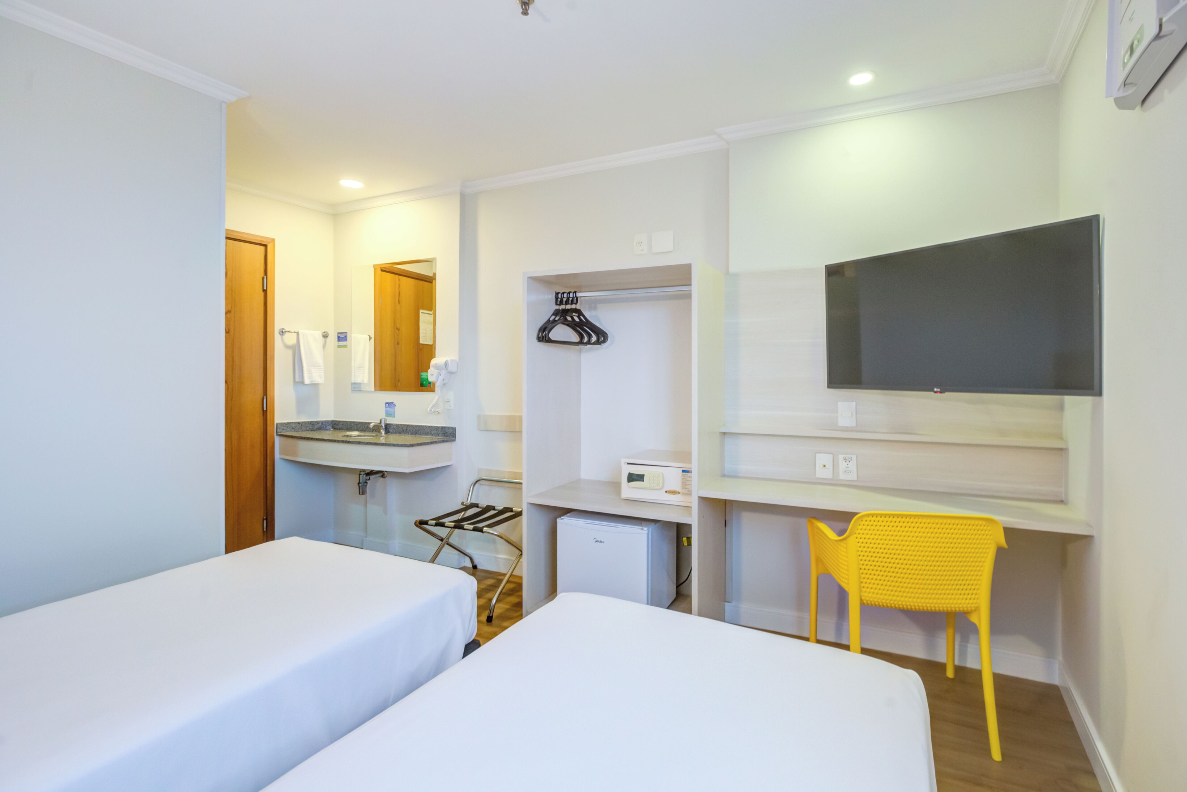 A hotel room with two beds, a desk, a TV, a small fridge, and a yellow chair.