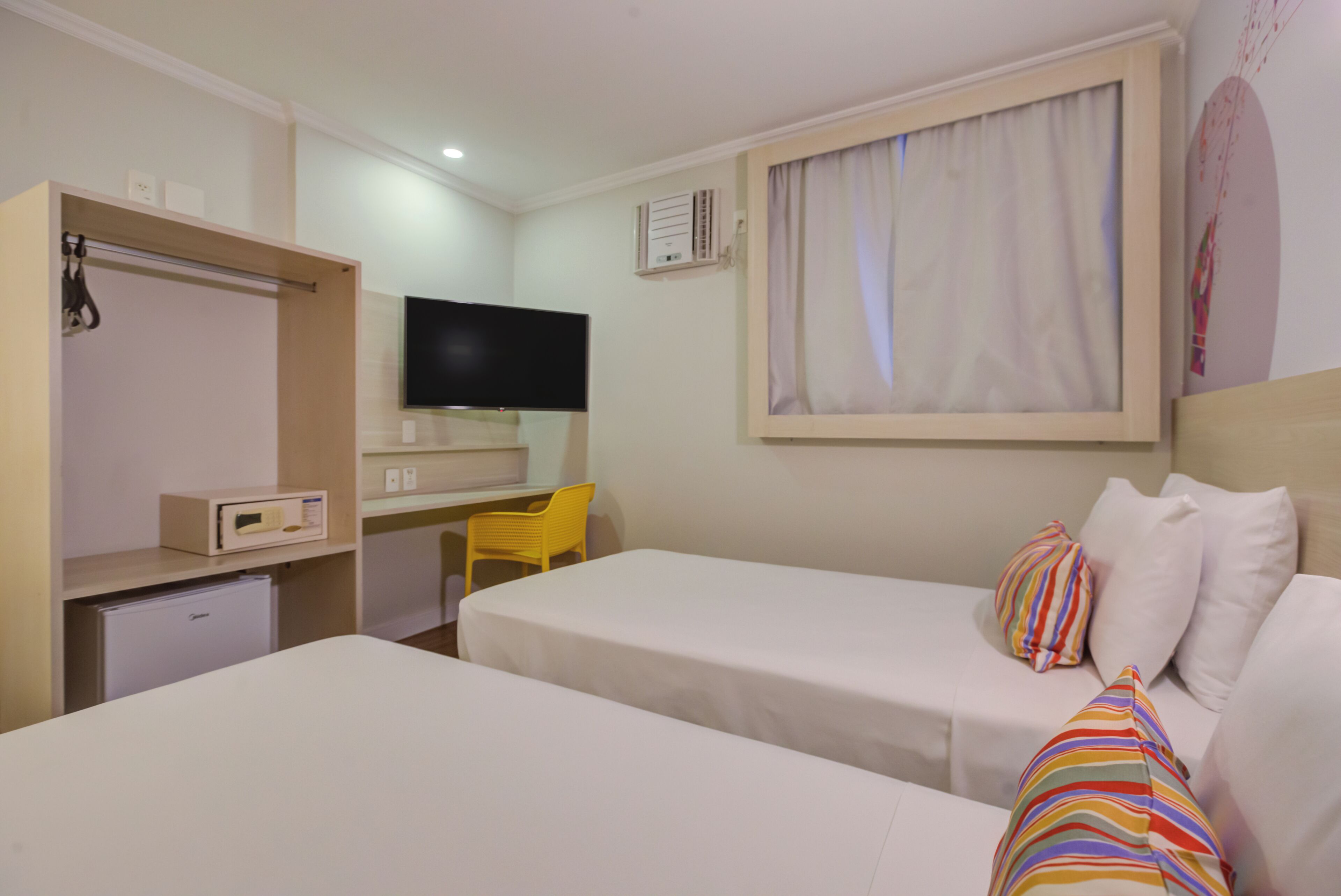 A hotel room with two beds, a desk, a TV, a small fridge, and a window with curtains.