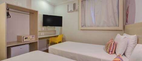 A hotel room with two beds, a desk, a TV, a small fridge, and a window with curtains.