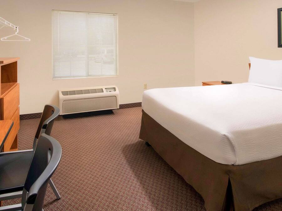 A hotel room with a bed, a desk, a chair, a heater, and a window with blinds.