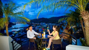 A couple seated at an outdoor table with a city view, overlooking a body of water and hills. - Royal Lotus Hotel Halong (Ha Long)