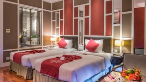 A hotel room with two beds, a large window with blinds, a bedside table with a lamp, and a small table with a fruit bowl. - Royal Lotus Hotel Halong (Ha Long)