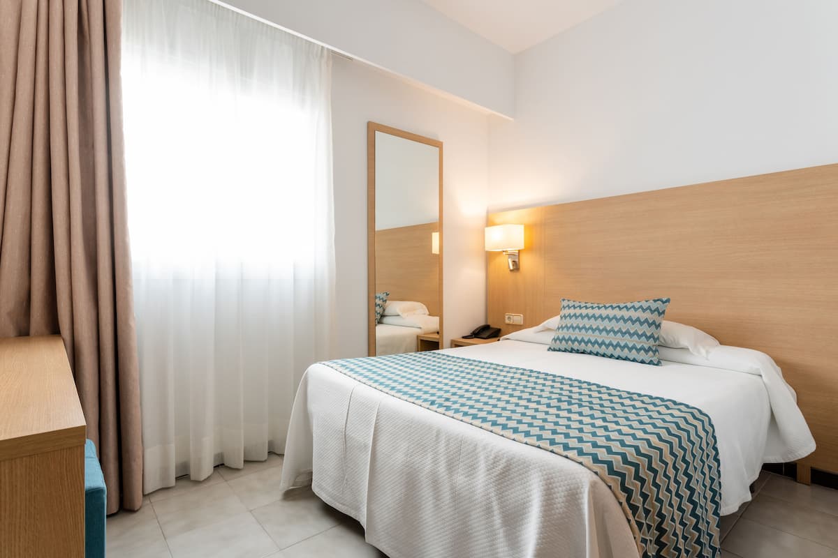 A hotel room with a bed, a nightstand, a mirror, a lamp, and a window with curtains.