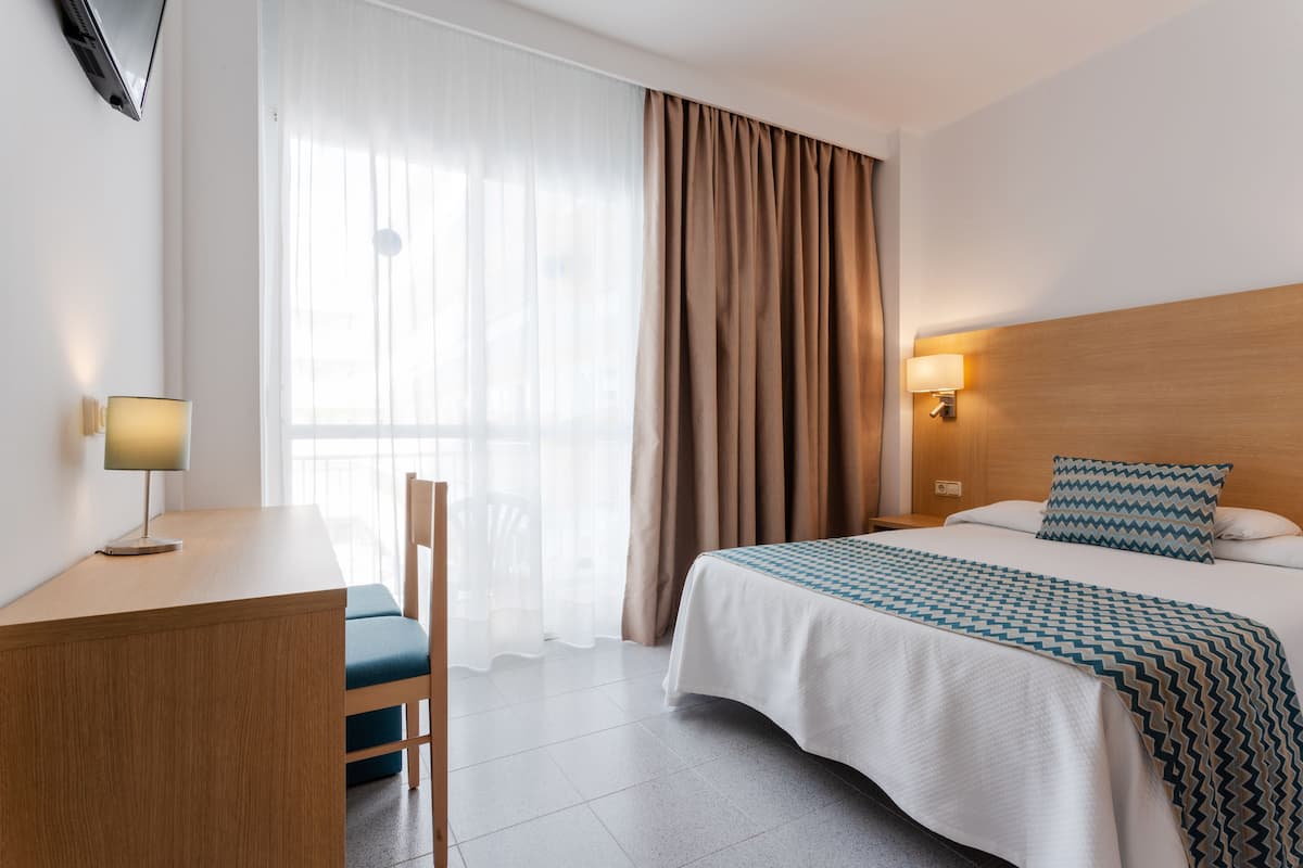 A hotel room with a bed, a desk, a chair, a lamp, and a window with curtains.