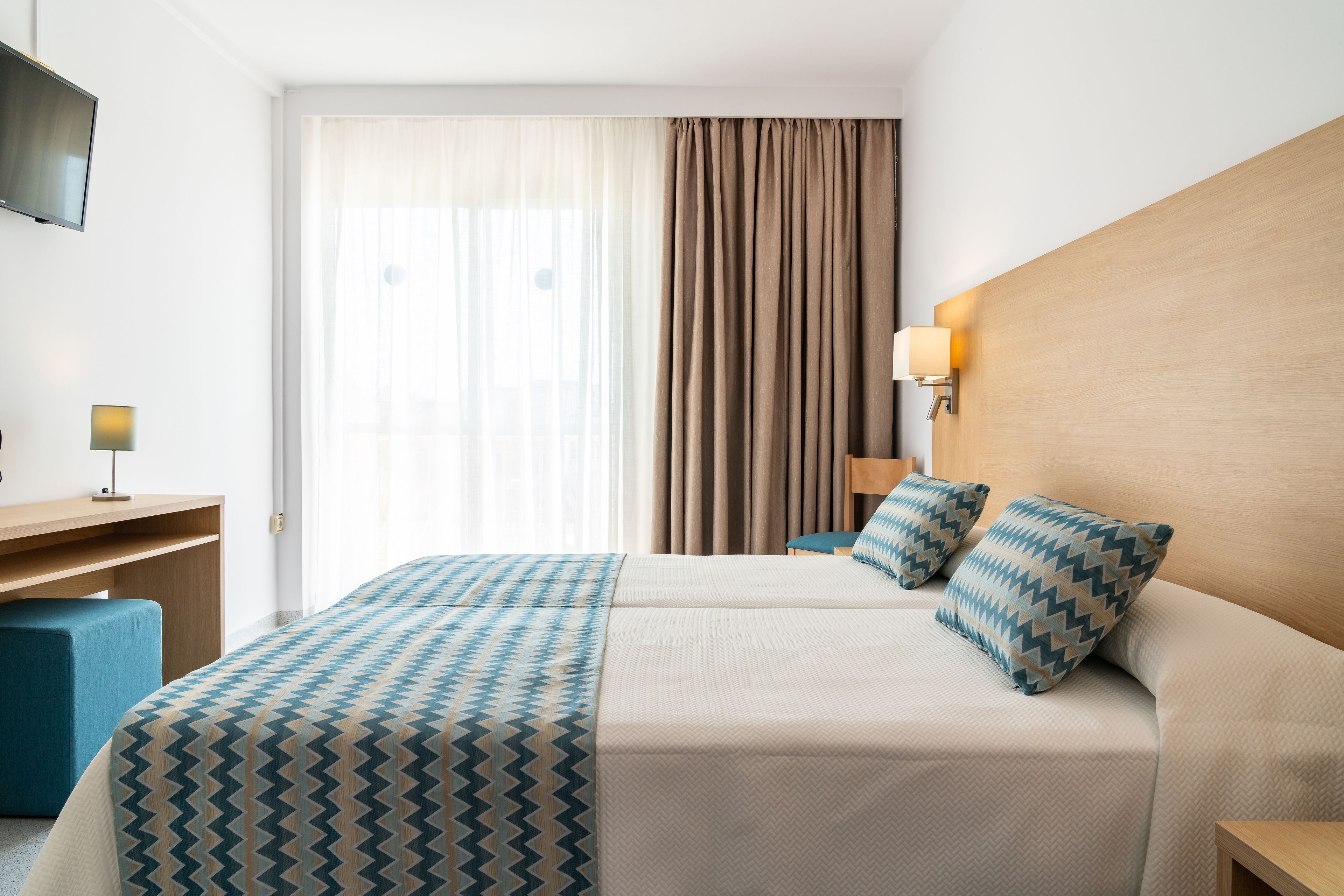 Una habitación de hotel con cama, escritorio, televisor y ventana con cortinas.