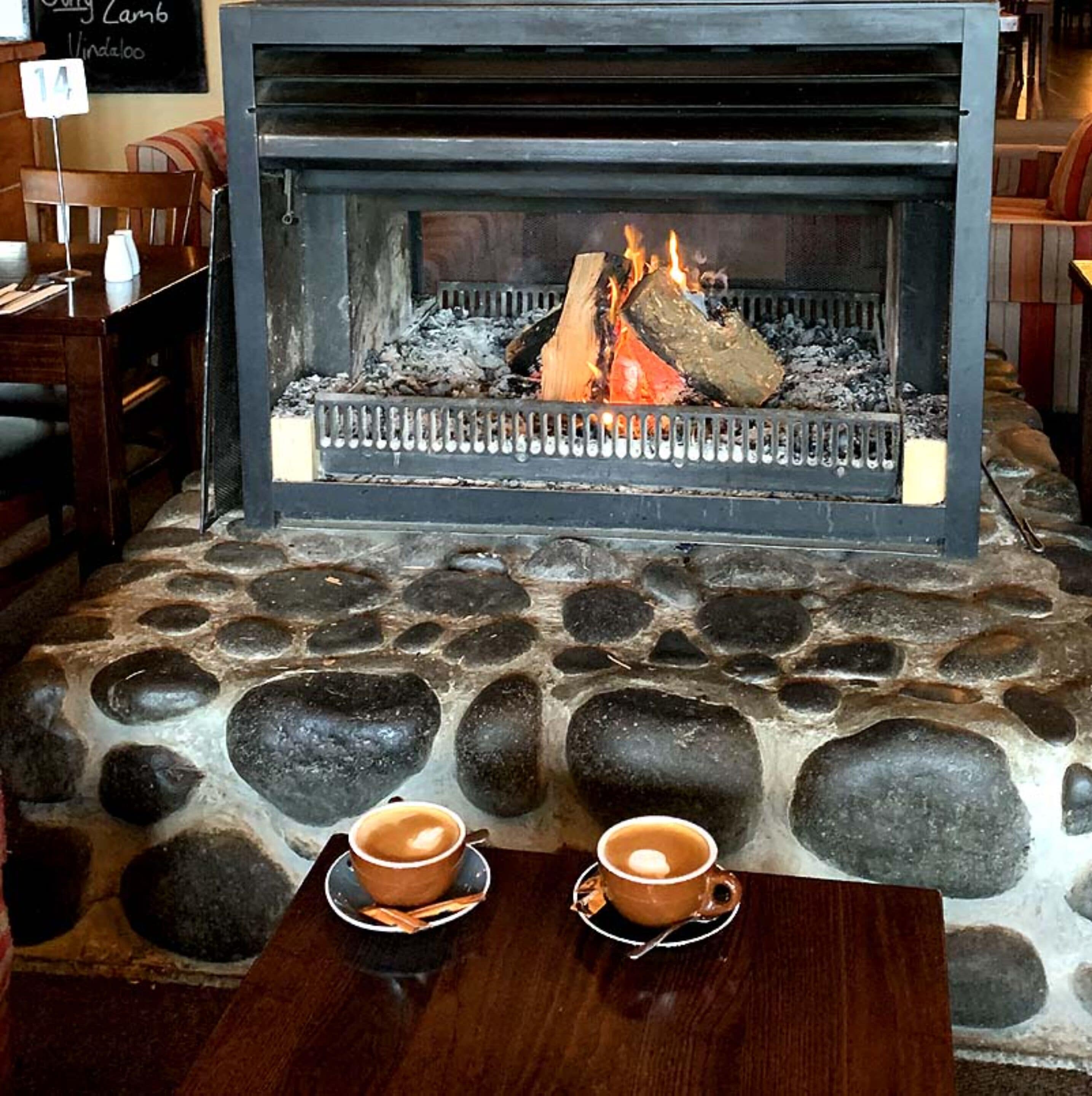 Lareira de pedra com fogo aceso, duas chávenas de café sobre uma mesa.