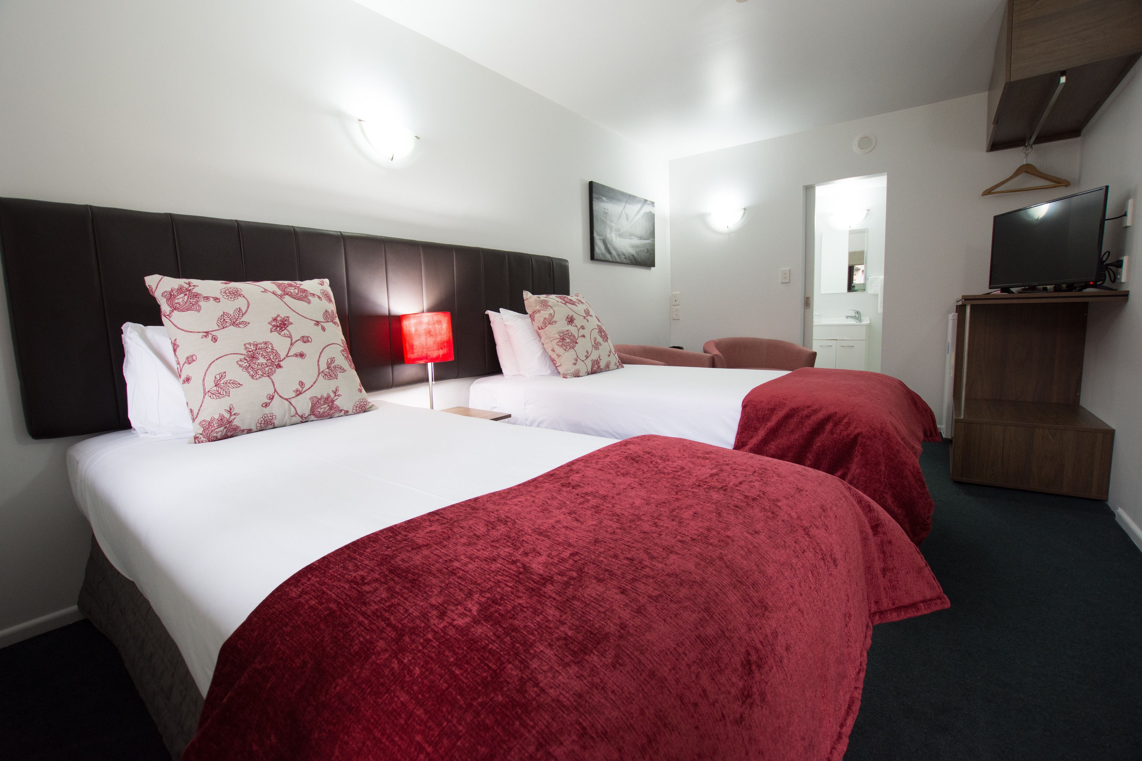 A hotel room with two beds, each with red and white bedding, a bedside table, a red lamp, a television on a wooden stand, and a mirror in the bathroom.