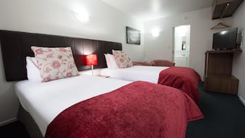 A hotel room with two beds, each with red and white bedding, a bedside table, a red lamp, a television on a wooden stand, and a mirror in the bathroom.