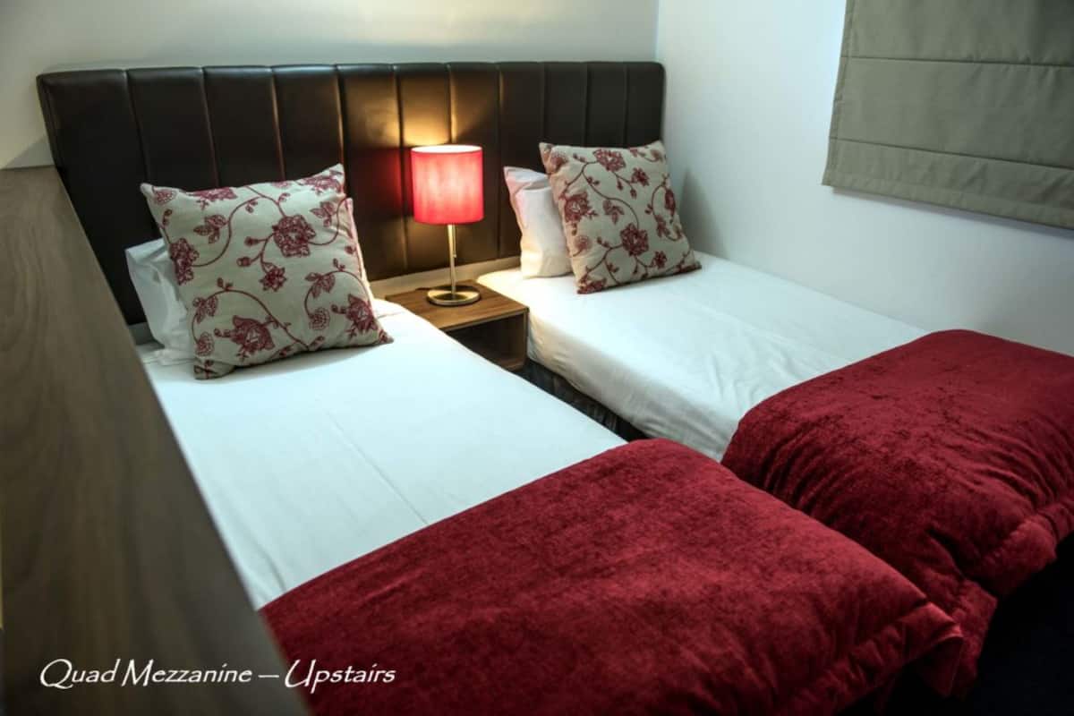 A hotel room with two single beds, a nightstand with a lamp, and a red blanket on one bed.