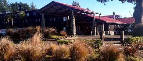 A lodge with a red roof and stone pillars, surrounded by a landscaped garden with tall grasses.