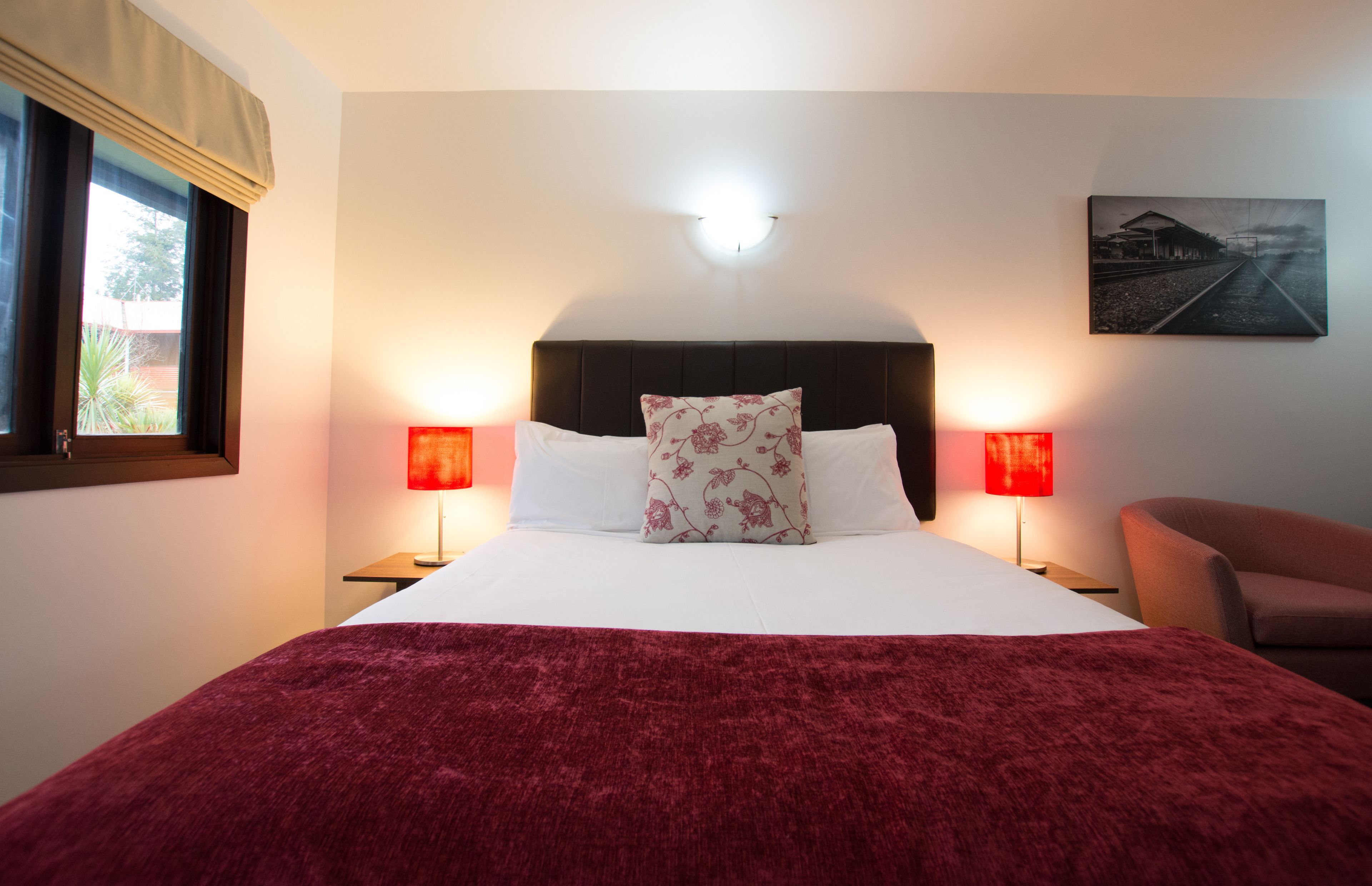 A hotel room with a large bed, two bedside lamps, a headboard, a red throw blanket, a chair, and a wall-mounted picture.