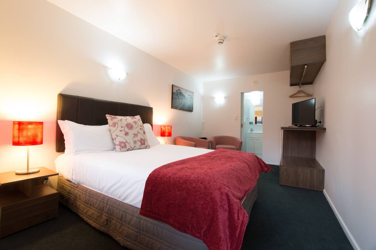 A hotel room with a bed, bedside lamps, a TV shelf, and a chair.
