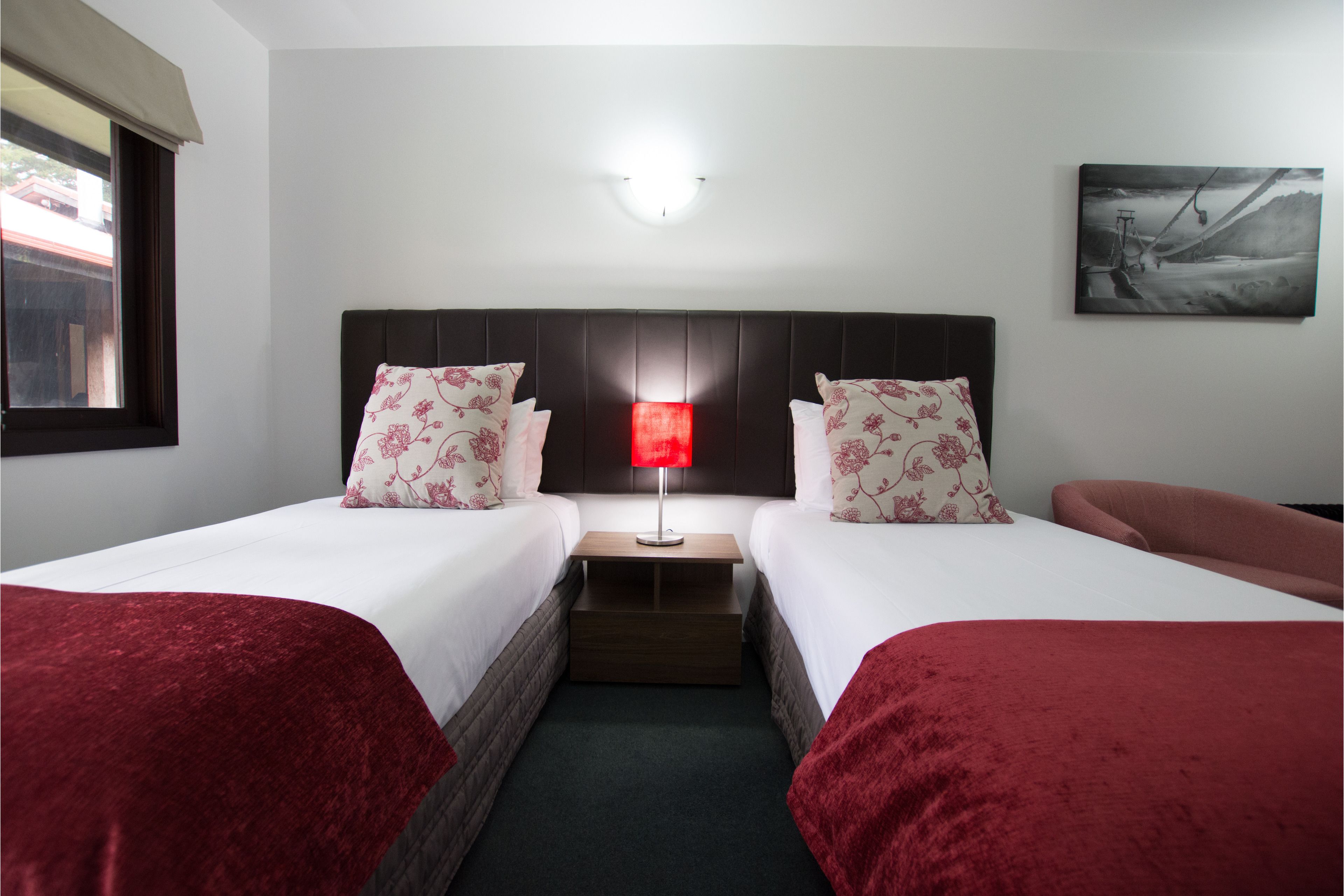 A hotel room with two beds, a nightstand with a lamp, a painting on the wall, and a window with a view of buildings.