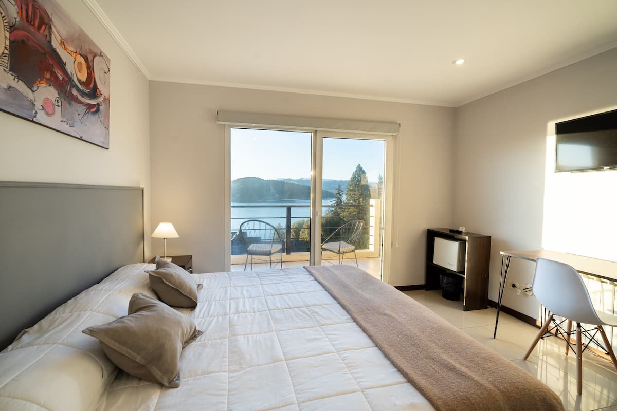 A bedroom with a large bed, a desk, a chair, and a view of a lake through a sliding door.