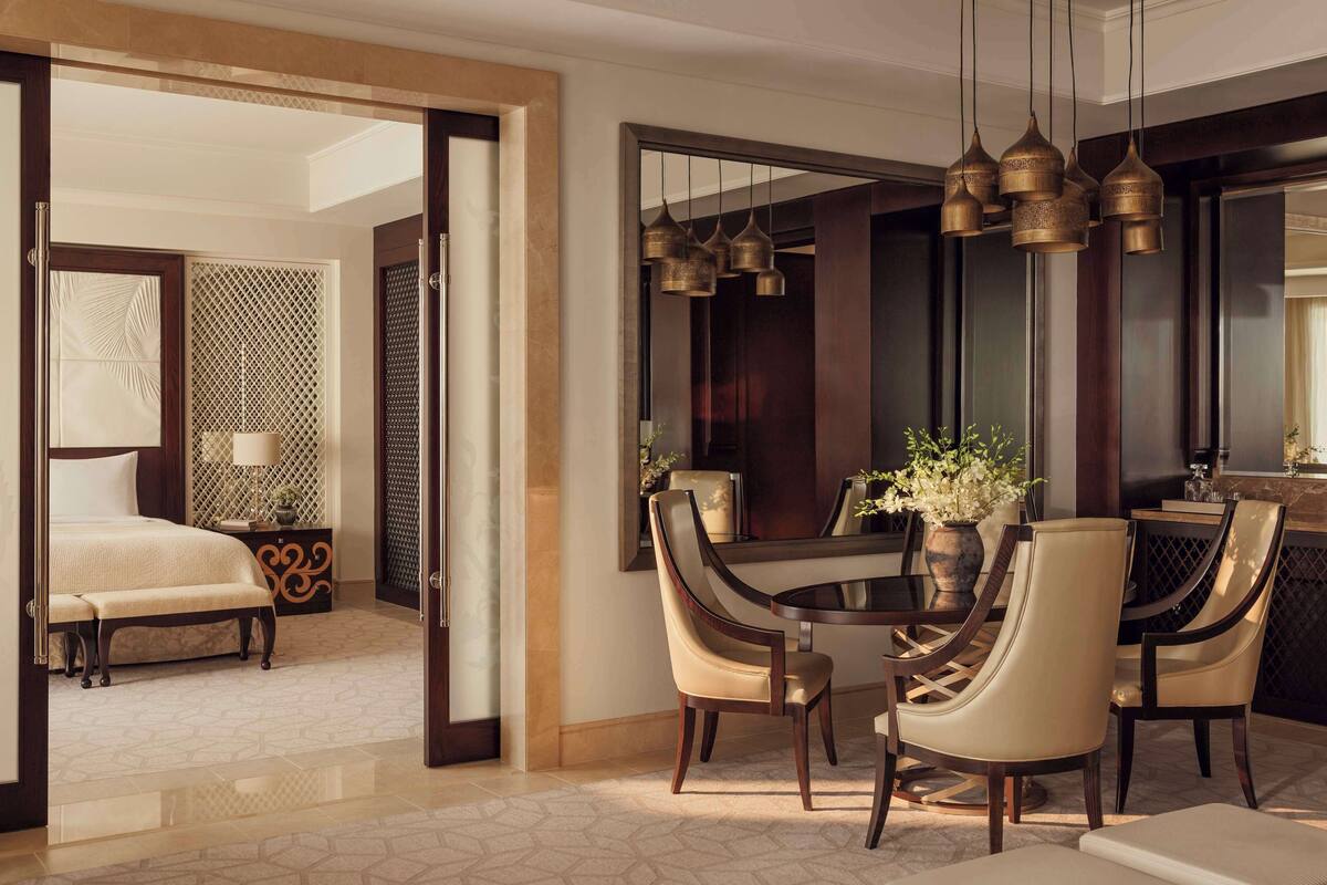 A modern hotel room with a large bed, a sitting area with a bench, a dining table with chairs, and a large mirror.