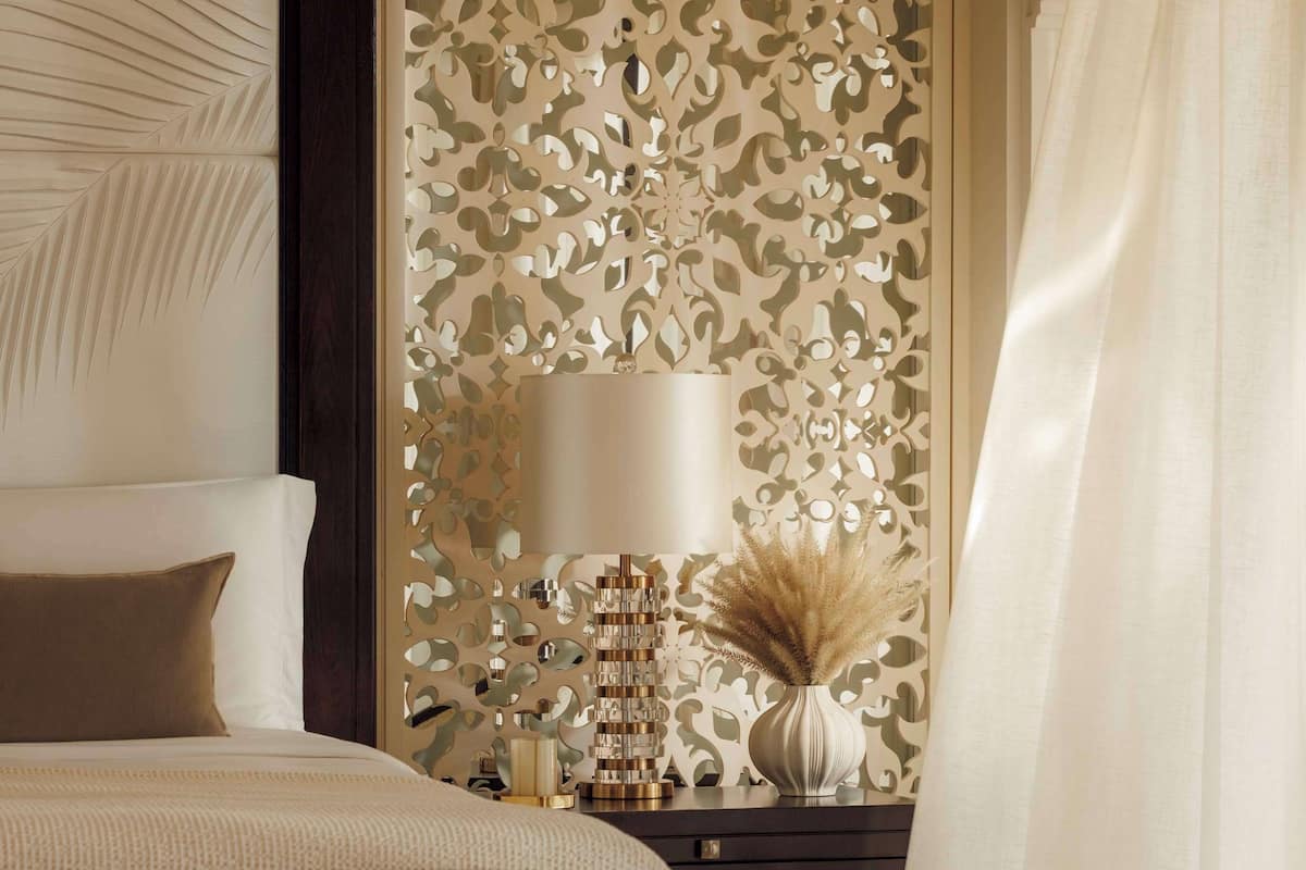 A bedroom with a bed, a bedside table with a lamp, a decorative wall, and a window with curtains.