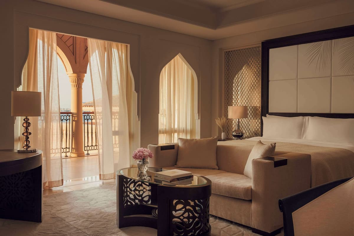 A modern living room with a sofa, a coffee table with a vase, and a lamp. There is a large window with curtains and a view of a balcony.
