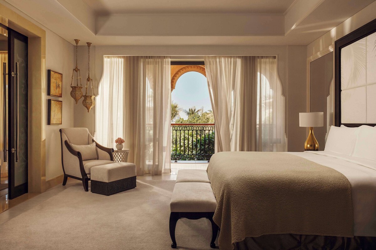 Een hotelkamer met een groot bed, een stoel, een bankje en uitzicht door een balkondeur.