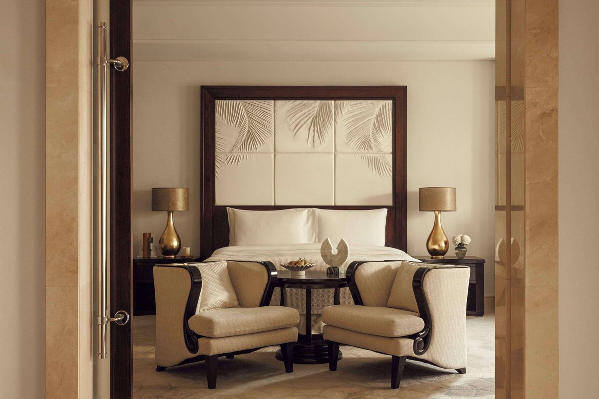 Een hotelkamer met een groot bed, twee fauteuils, een klein tafeltje en een decoratief hoofdeinde.