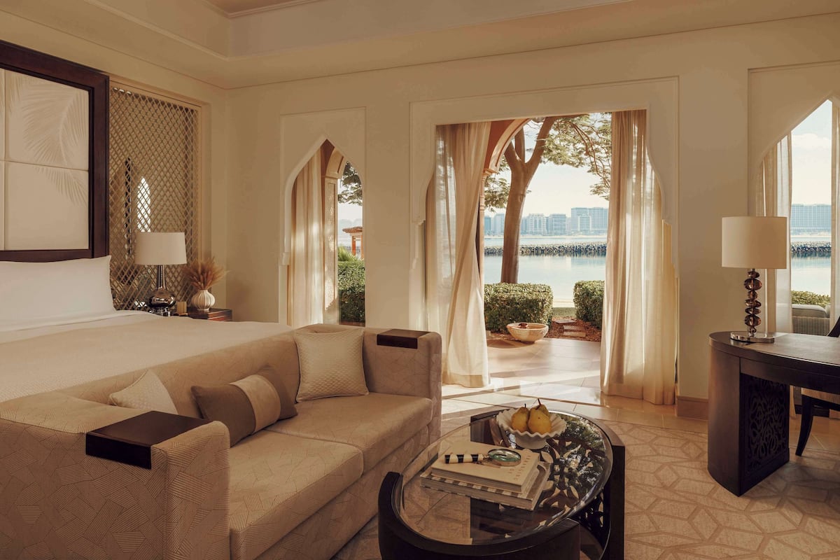 A hotel room with a bed, a sofa, a coffee table with fruits, and a view of a body of water.