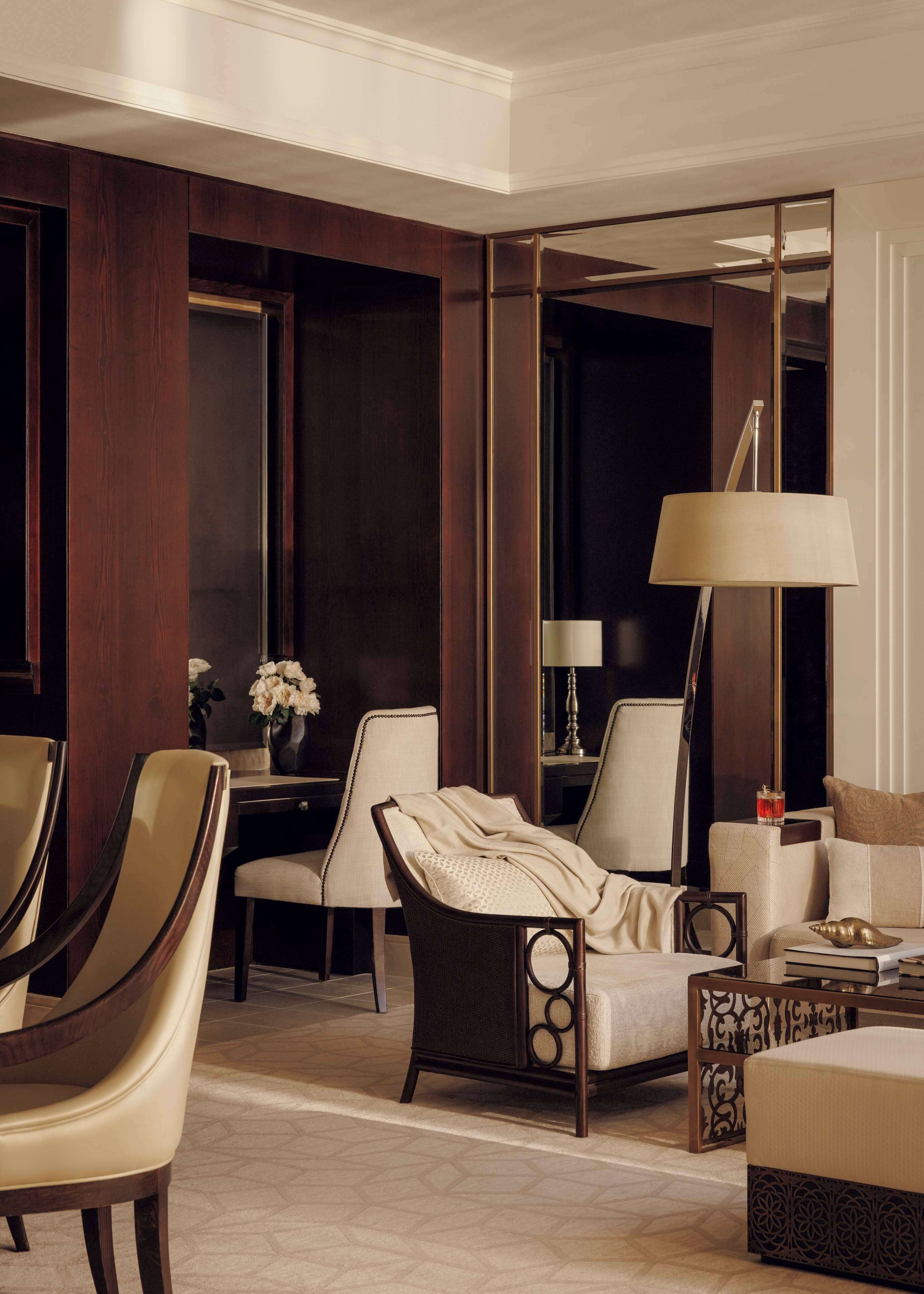 Una habitación de hotel moderna con paneles de madera oscura, un espejo grande, un sofá, dos sillones, una mesita con un jarrón de flores y una lámpara de pie.