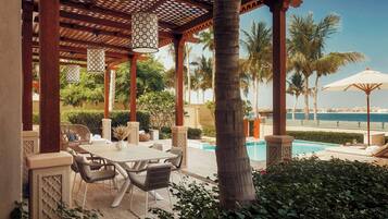 A covered outdoor area with a pool, palm trees, and a view of the sea.