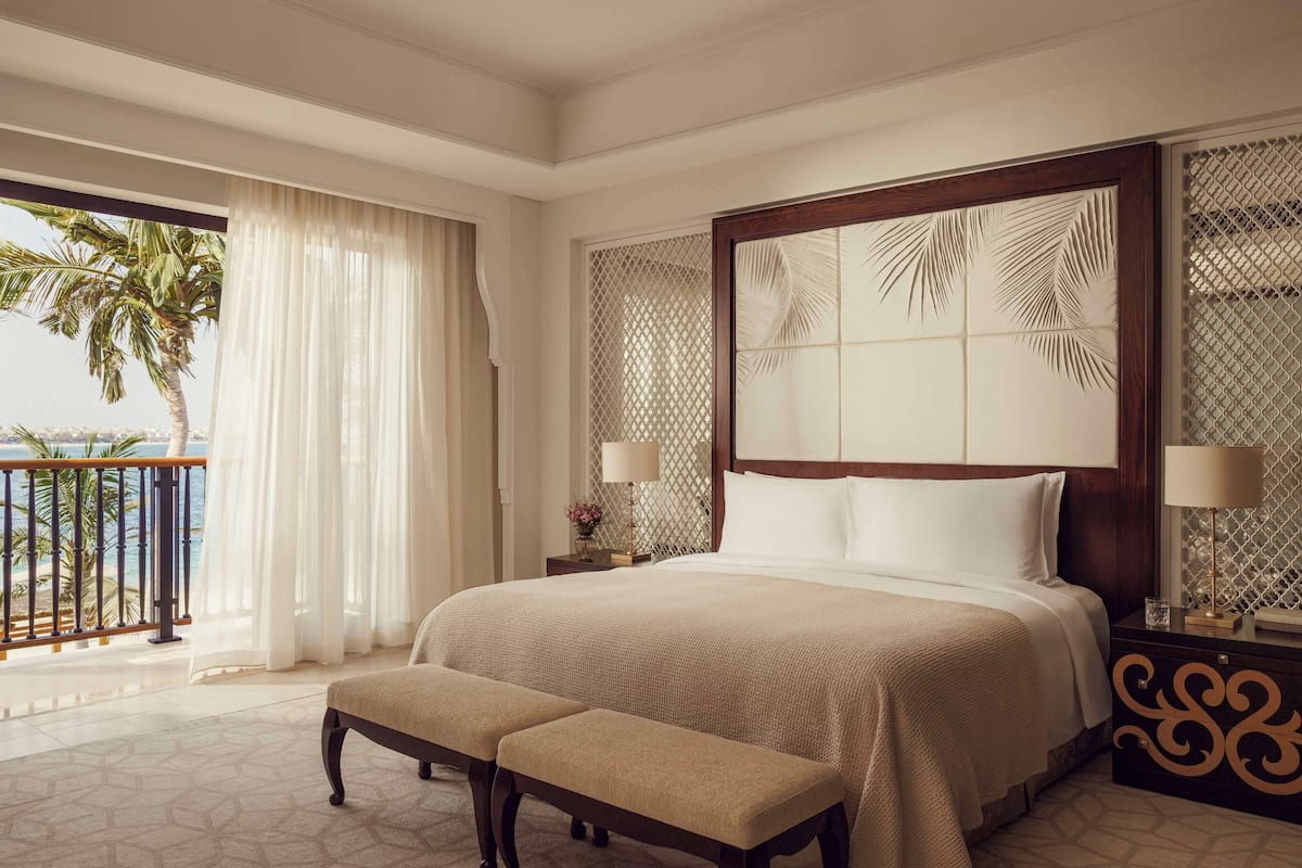 A hotel room with a large bed, a bench, a nightstand with lamps, and a view of palm trees and the sea.