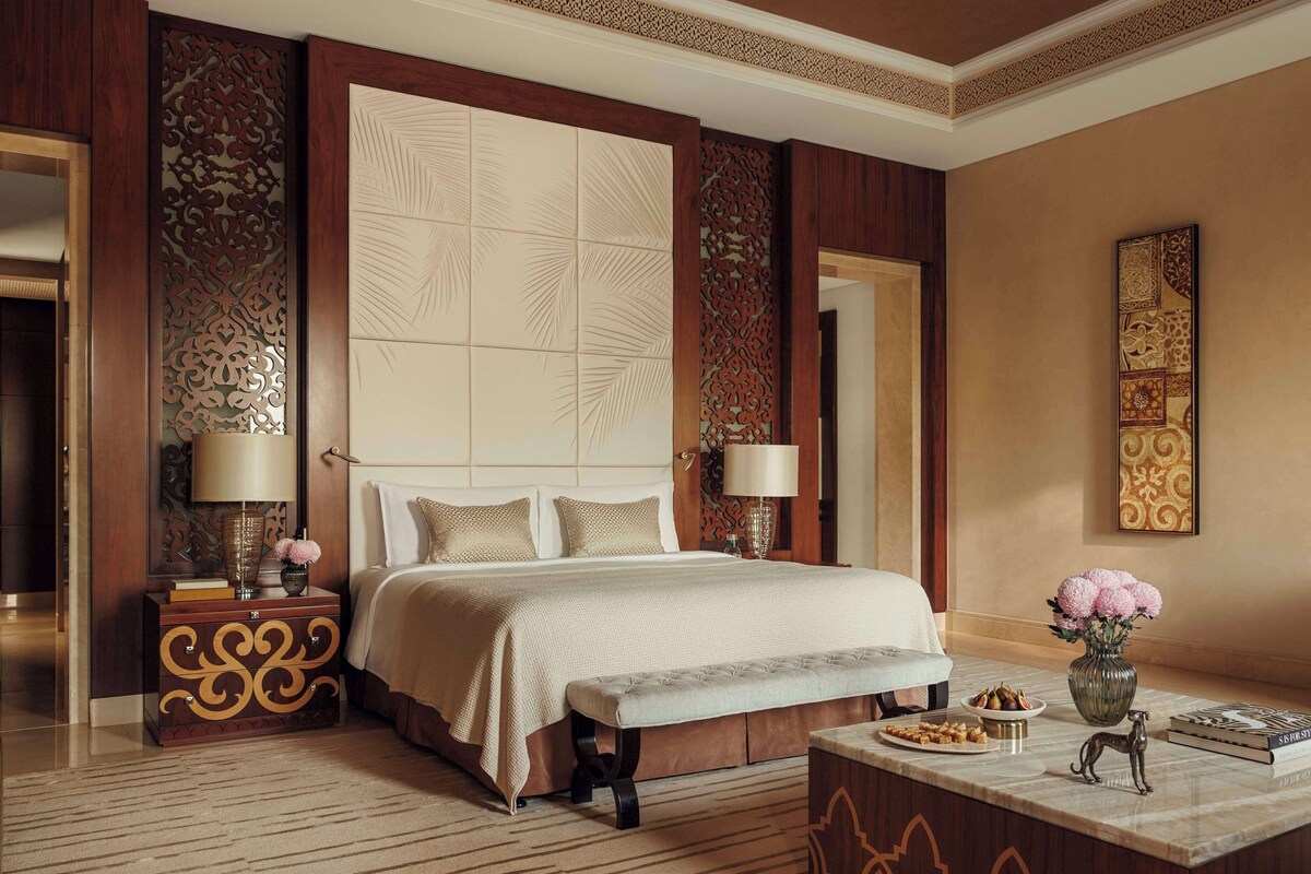 Een hotelkamer met een groot bed, twee nachtkastjes met lampen, een bankje, een decoratieve vaas met bloemen en een ingelijste kunstwerk aan de muur.