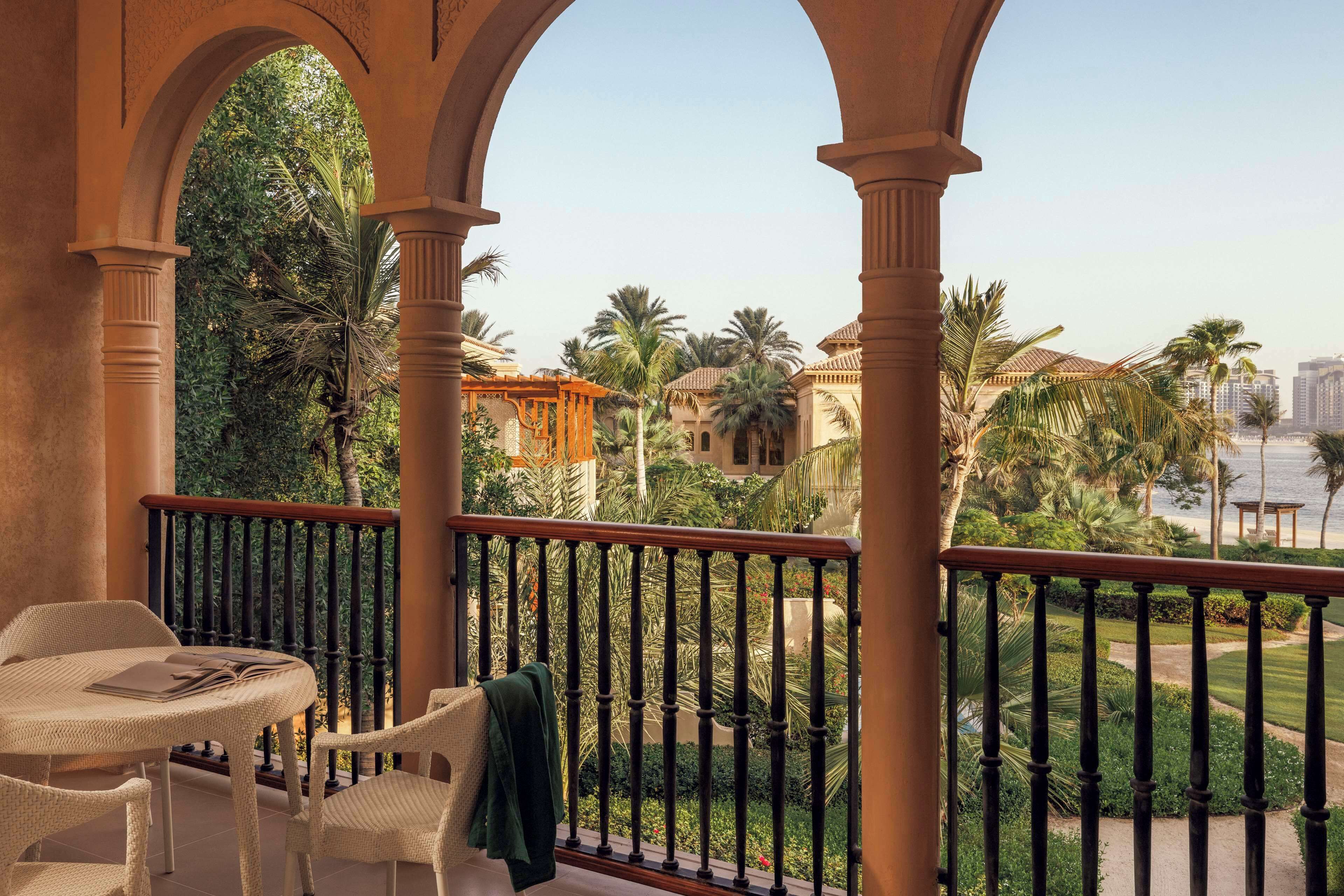 Ein Balkon mit Tisch und Stühlen, umgeben von Palmen und Blick auf eine Stadtlandschaft.