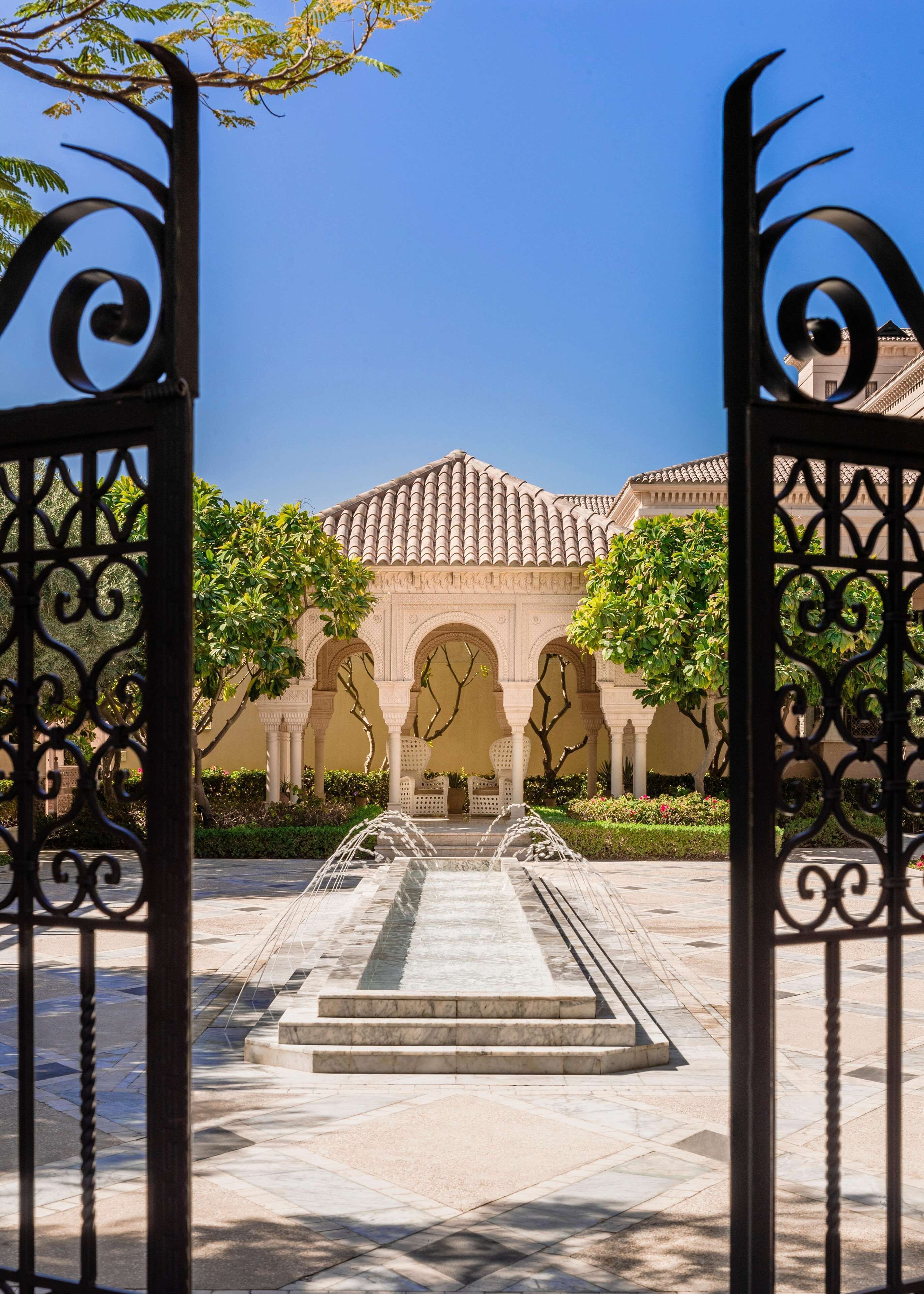 An entrance to a courtyard with a fountain, surrounded by arches and a building with a tiled roof.