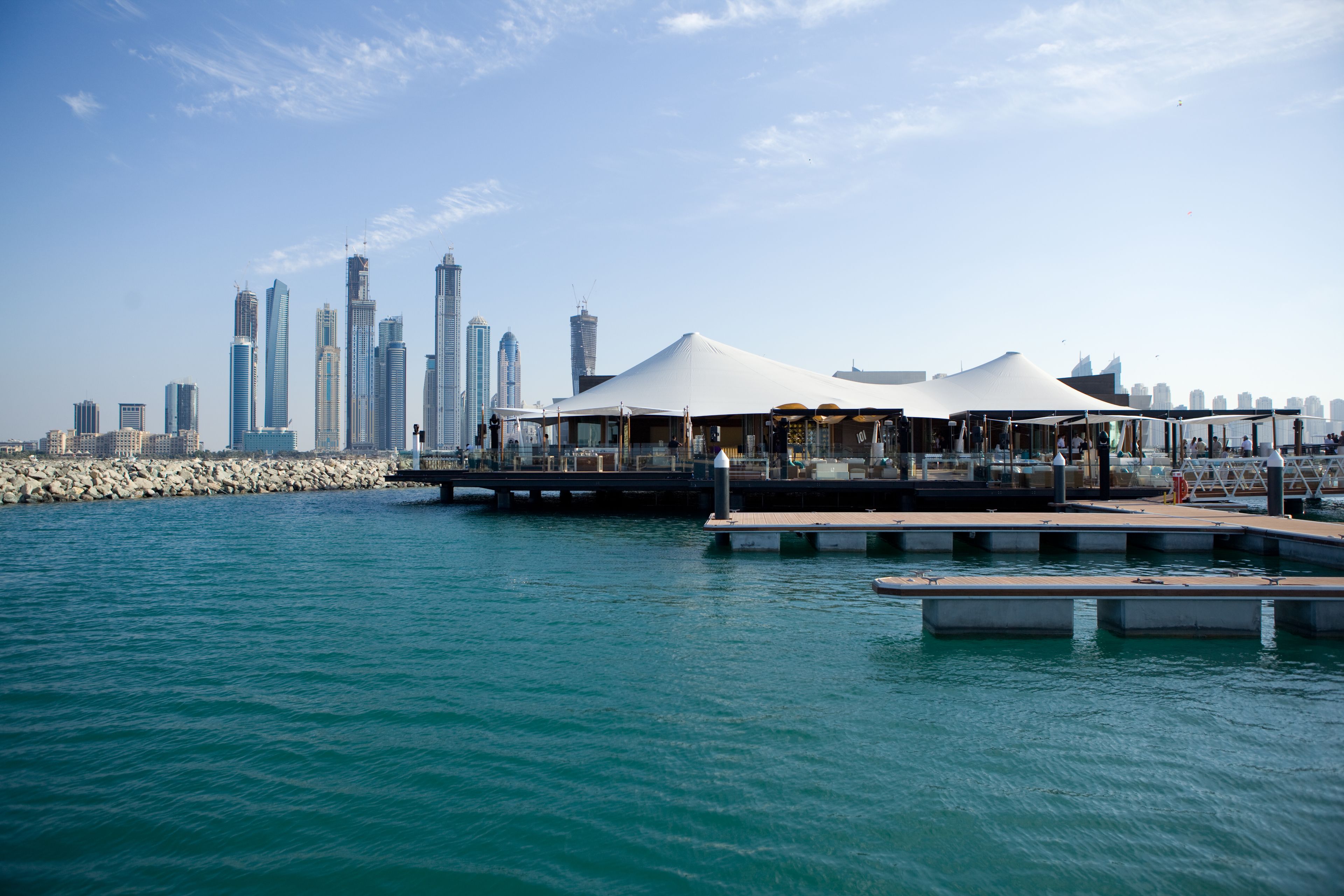 A waterfront area with a row of white tents, a dock, and a city skyline in the background.