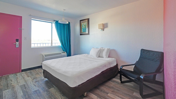 Una camera da letto con un letto, una sedia, una finestra con tende, un quadro appeso al muro e una porta rosa.