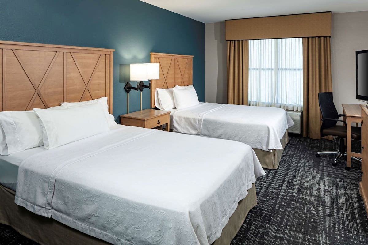 A hotel room with two beds, a desk, a chair, a TV, and a window with curtains.