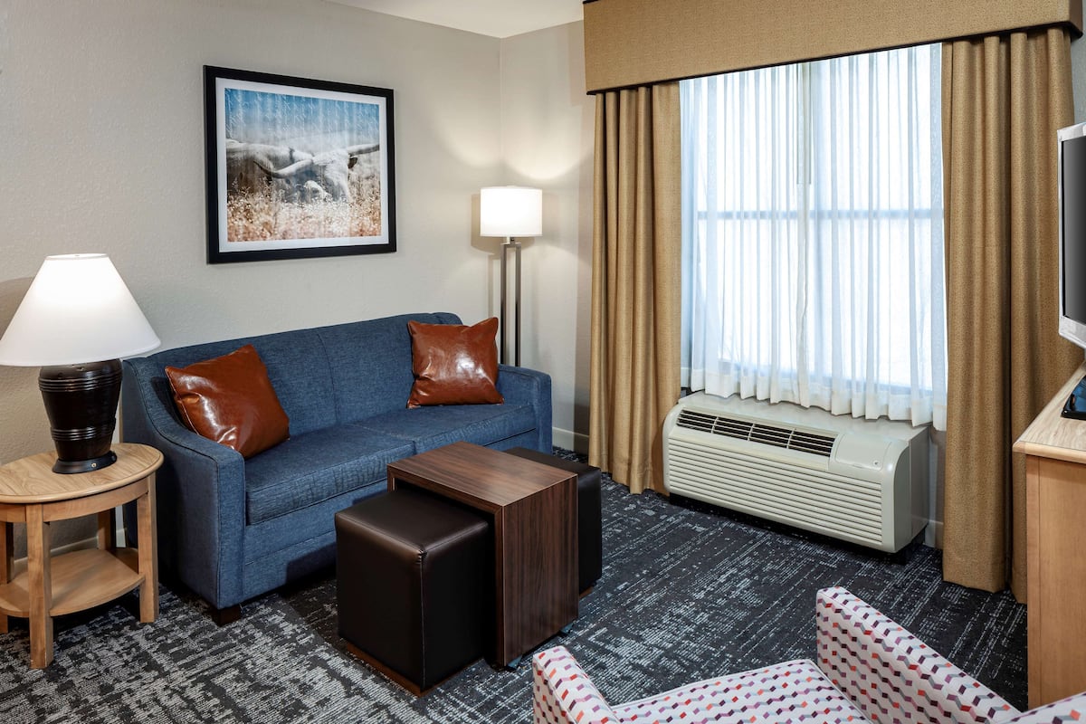 A hotel room with a blue sofa, a wooden side table with a lamp, a patterned armchair, a large window with curtains, and a framed picture on the wall.