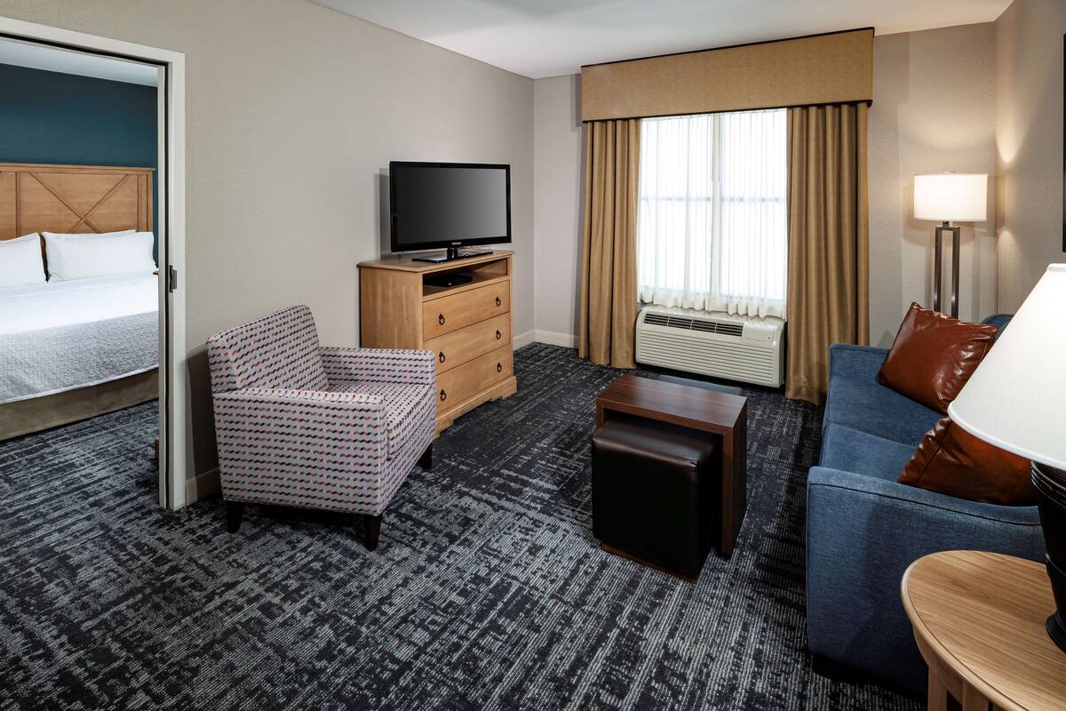 A hotel room with a bed, a TV on a wooden dresser, a blue sofa, a patterned armchair, a small table, and a lamp.