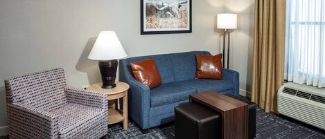 Habitación de hotel con un sofá azul, un sillón estampado, una mesita y un cuadro en la pared.