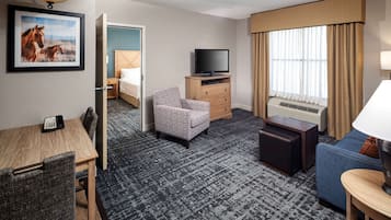 Quarto de hotel com cama, televisão, escrivaninha com telefone, poltrona estampada, sofá azul e janela com cortinas.
