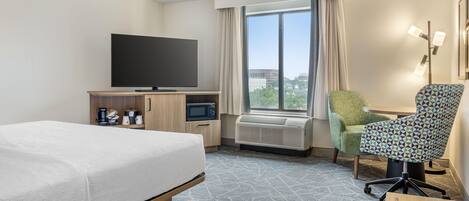 A hotel room with a bed, a desk with a flat-screen TV, a chair, a coffee maker, and an air conditioner.