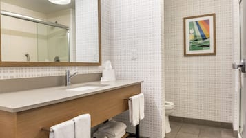 A modern bathroom with a large mirror, a sink, and a toilet. There is a towel rack with towels and a small shelf with a bottle.