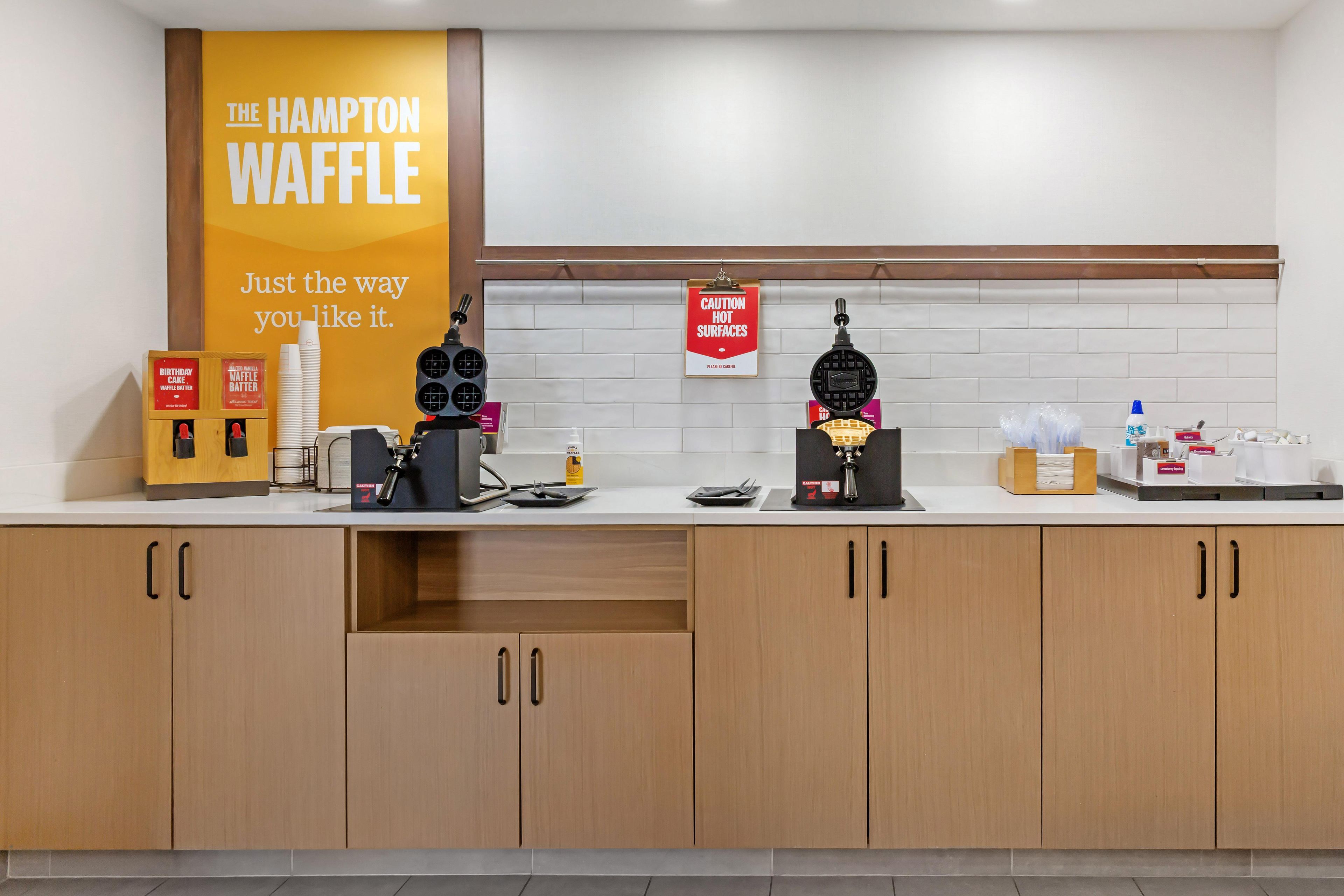 A kitchen counter with waffle-making equipment, a sign for caution hot surfaces, and a birthday cake box.