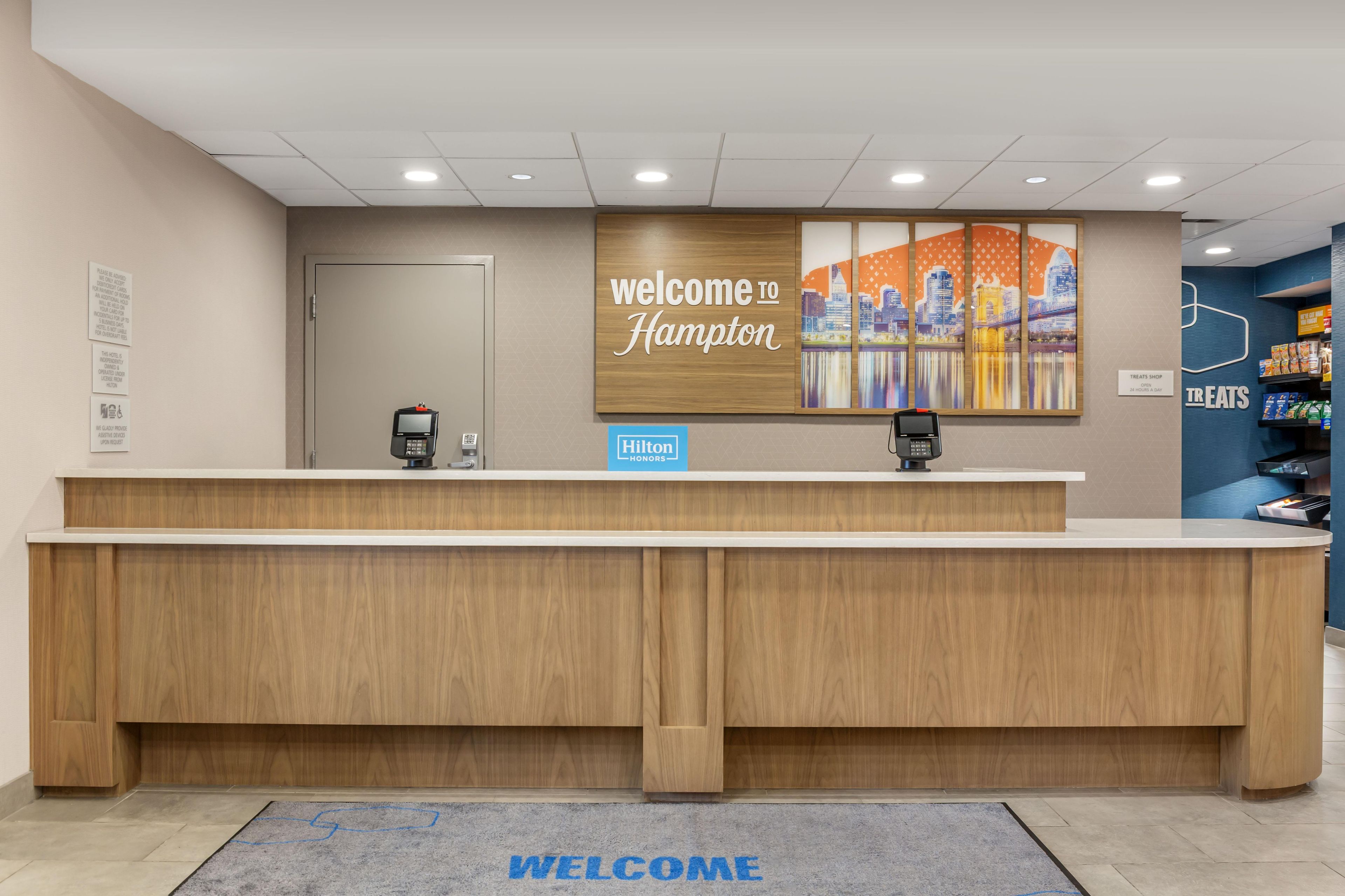 A hotel reception desk with a 'Welcome to Hampton' sign, a digital kiosk, and a 'Treats Shop' sign.